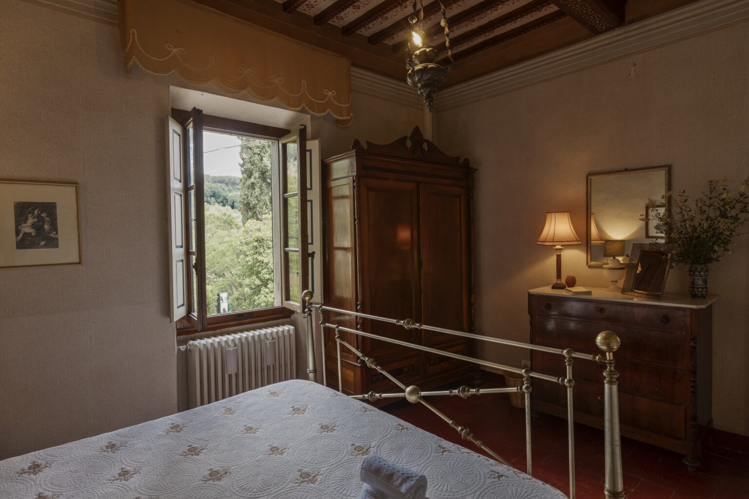 Cozy bedroom interior featuring a vintage wooden wardrobe, a dresser with a lamp, and a window with scenic views. The room is decorated in soft tones with a floral-patterned bedspread and warm lighting. | Only on SopranoVillas