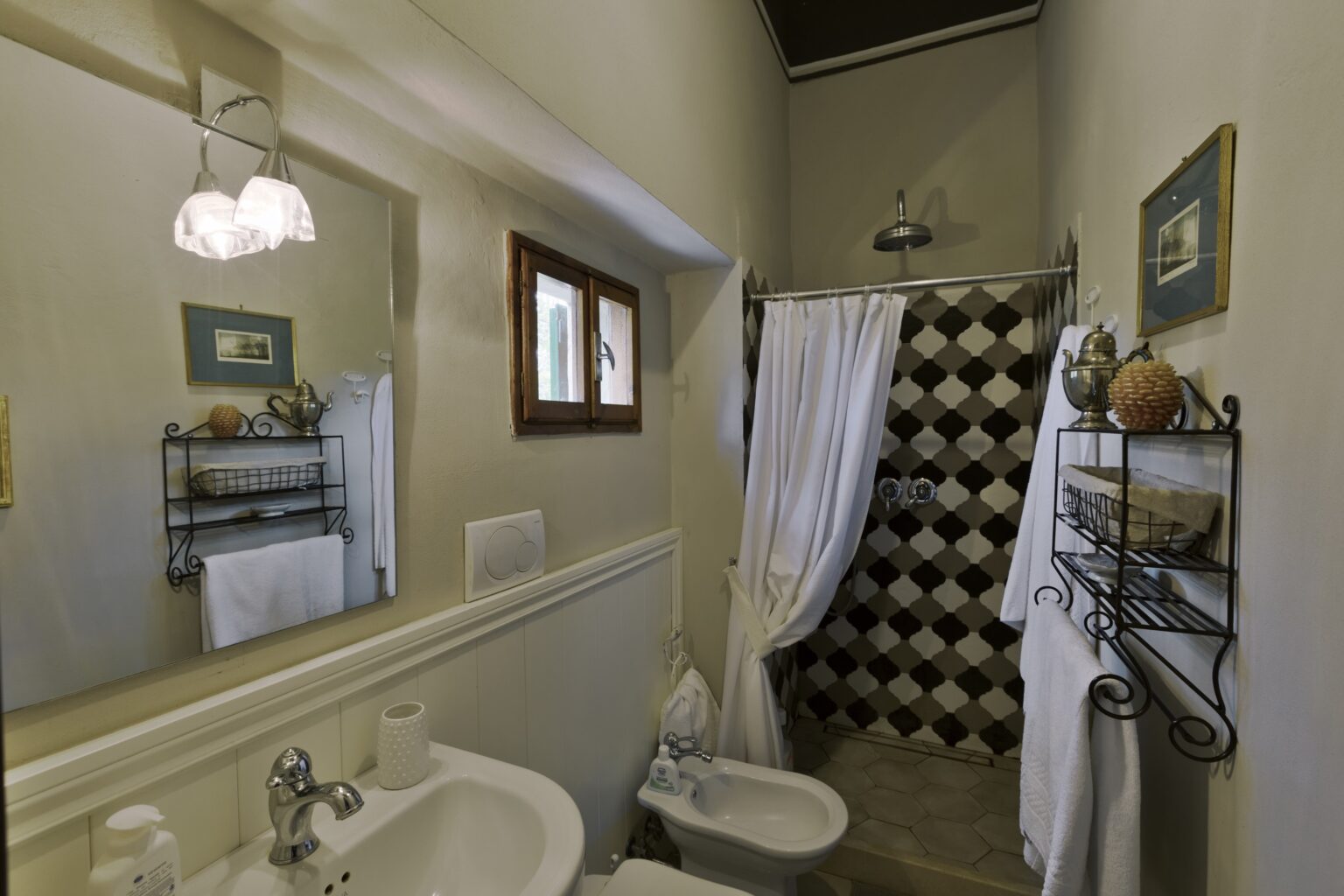 Modern bathroom featuring a shower with a black and white tiled wall, a bidet, and a stylish sink. Decorative shelves hold towels and toiletries, with natural light coming from a small window. | Only on SopranoVillas