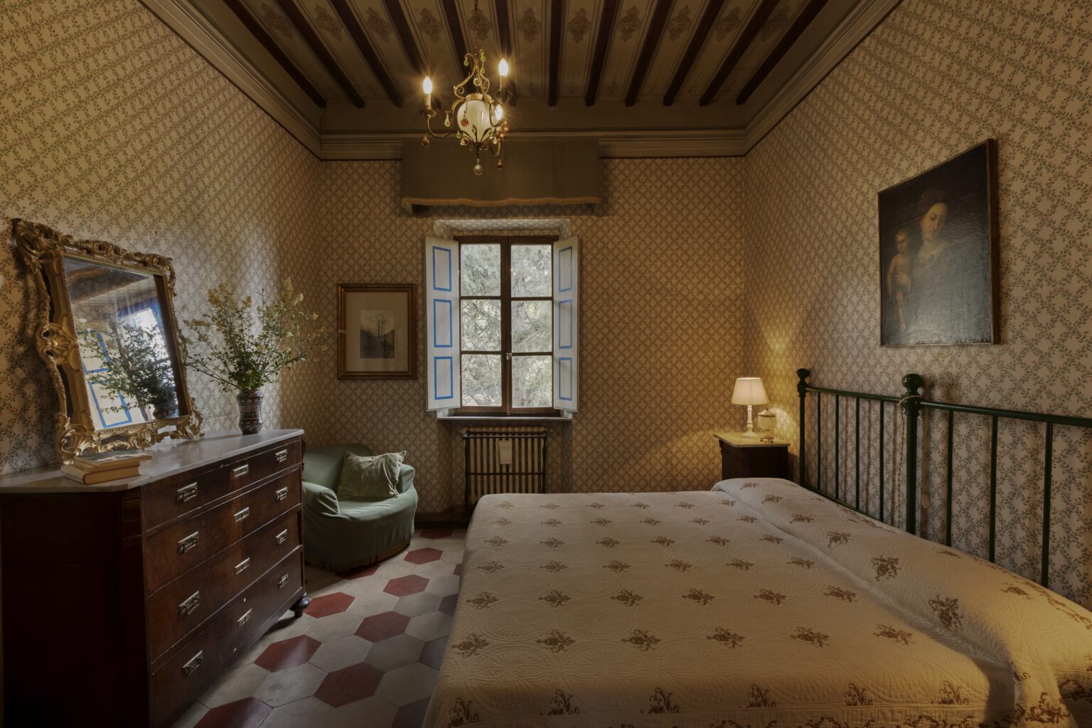 Cozy bedroom featuring a double bed with a patterned bedspread, a vintage wooden dresser, a green armchair, and natural light streaming through a large window. The room has decorative wallpaper and a chandelier, creating a warm and inviting atmosphere. | Only on SopranoVillas