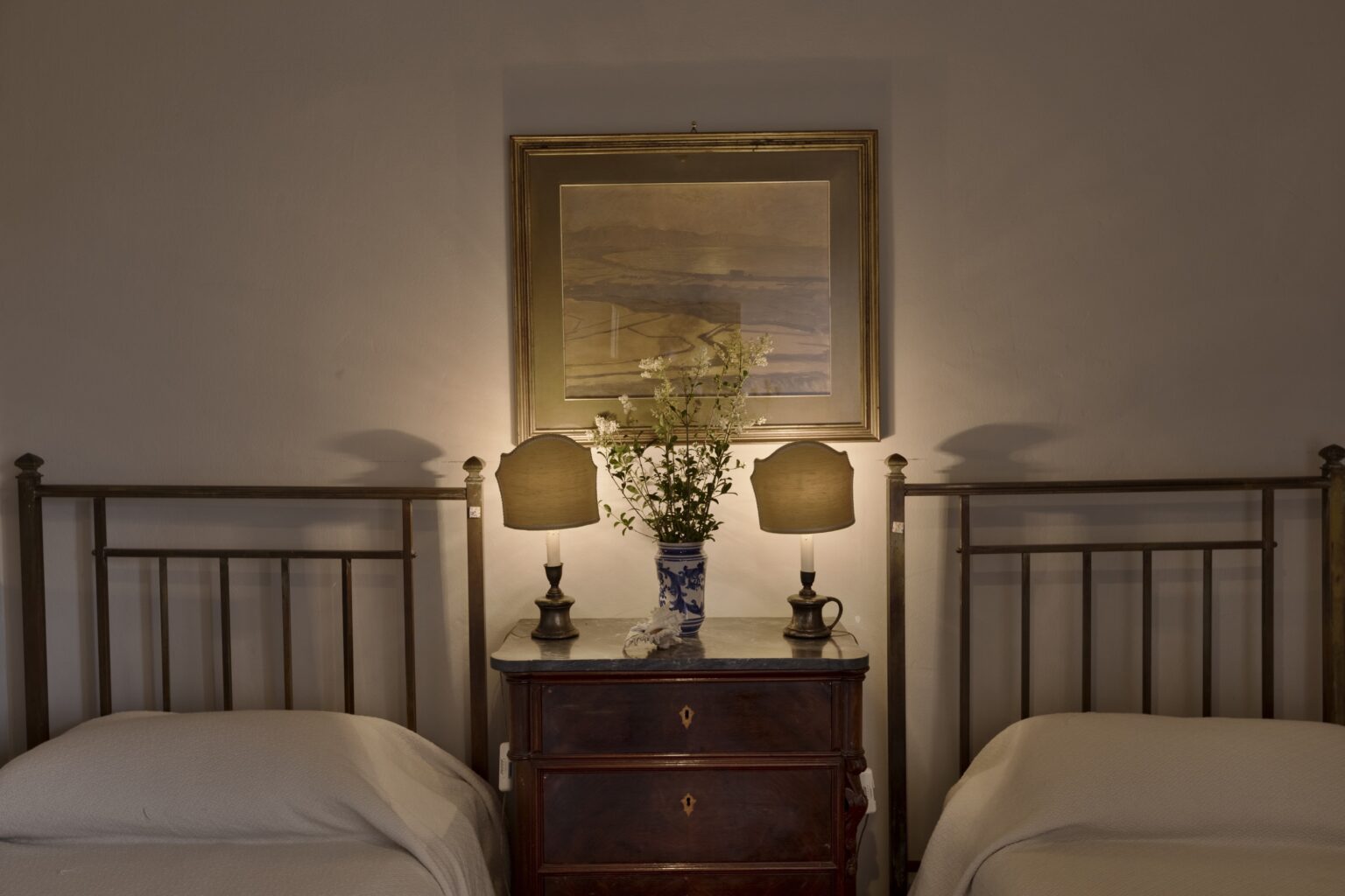 Cozy bedroom interior featuring two twin beds with soft bedding, a wooden nightstand with a marble top, and two elegant table lamps. A floral arrangement in a decorative vase and a framed landscape painting add charm to the serene atmosphere. Perfect for a relaxing getaway. | Only on SopranoVillas