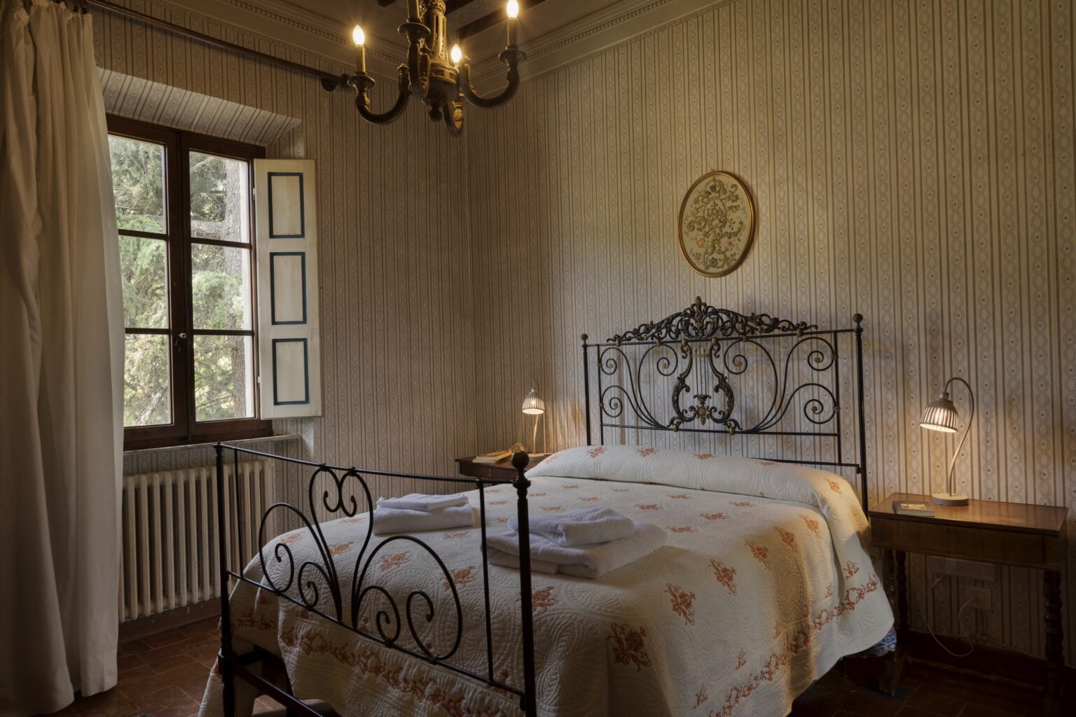 Cozy bedroom featuring an iron bed frame, soft bedding, and warm lighting from bedside lamps, with a vintage chandelier and a large window allowing natural light. | Only on SopranoVillas