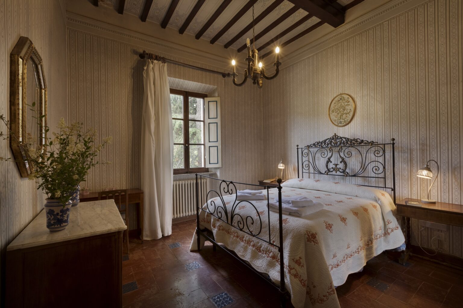 Cozy bedroom featuring a wrought iron bed, wooden beams, and a vintage chandelier, with natural light streaming through a window and soft decor elements. | Only on SopranoVillas