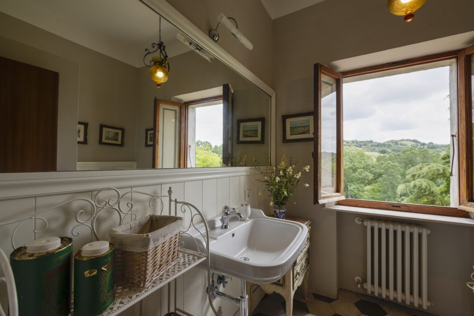 Bright and airy bathroom featuring a vintage sink, decorative storage baskets, and a large window with scenic views of greenery. Natural light illuminates the space, enhancing the charming decor and inviting atmosphere. | Only on SopranoVillas
