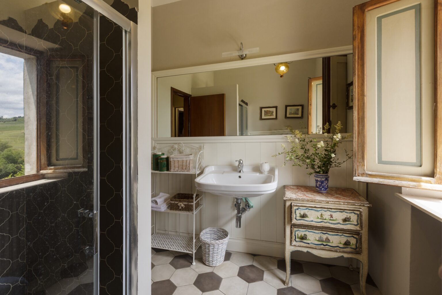 Modern bathroom interior featuring a shower, sink, and vintage-style furniture, with natural light streaming through windows and decorative elements like flowers and artwork. | Only on SopranoVillas