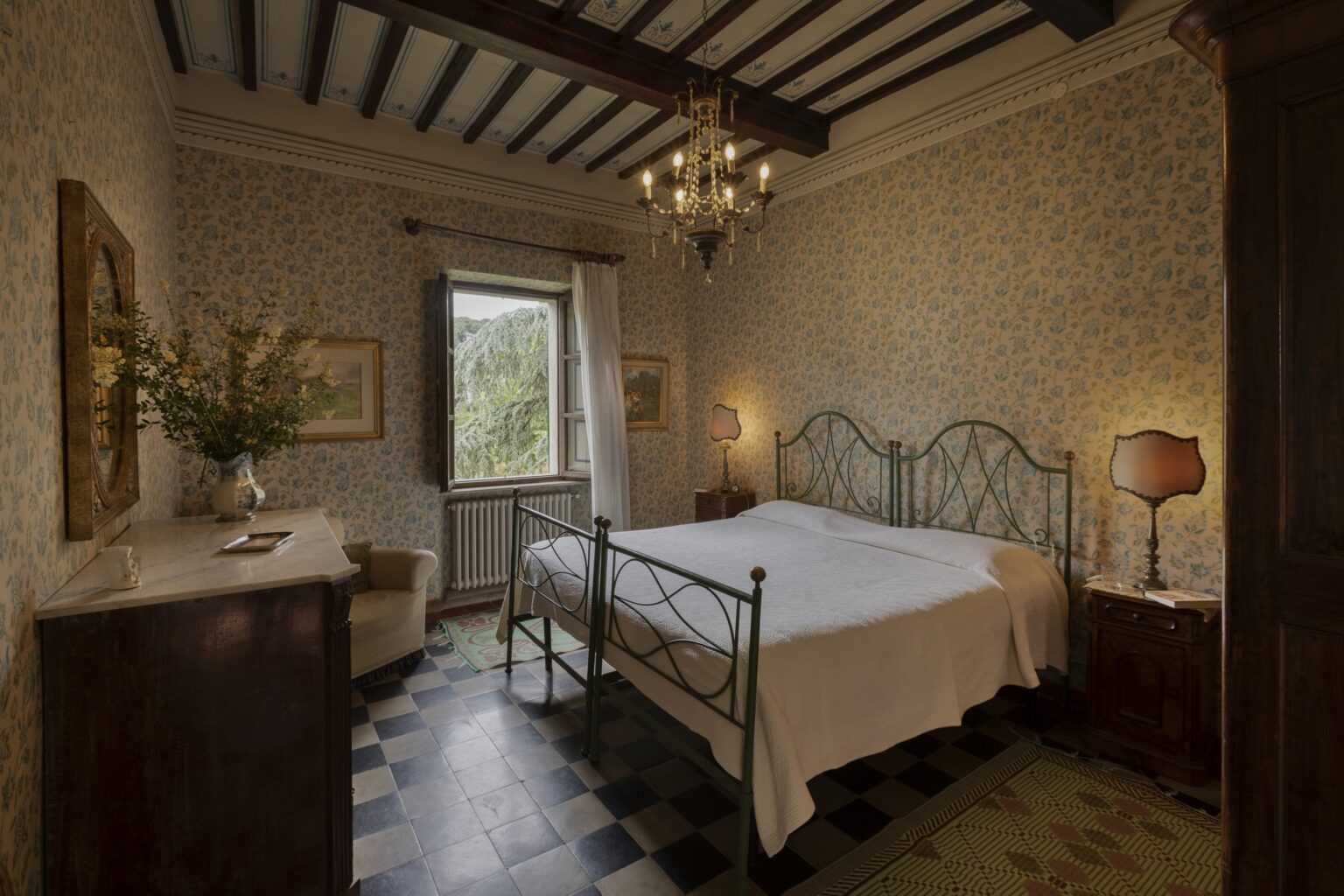 Cozy bedroom featuring a vintage metal bed frame, floral wallpaper, a chandelier, and a wooden dresser, with natural light streaming through a window. | Only on SopranoVillas
