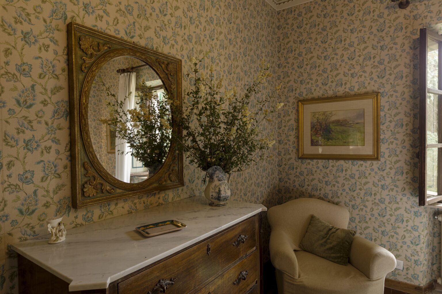 Cozy interior of a vintage-style room featuring floral wallpaper, a round mirror above a wooden dresser, a vase with greenery, and a plush armchair, creating a warm and inviting atmosphere. | Only on SopranoVillas