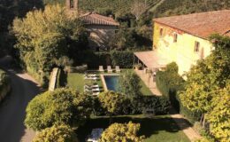 Scenic view of a Tuscan villa with a swimming pool, surrounded by lush gardens and cypress trees, featuring a long dining table set for an outdoor meal, with vineyards in the background under a clear blue sky.