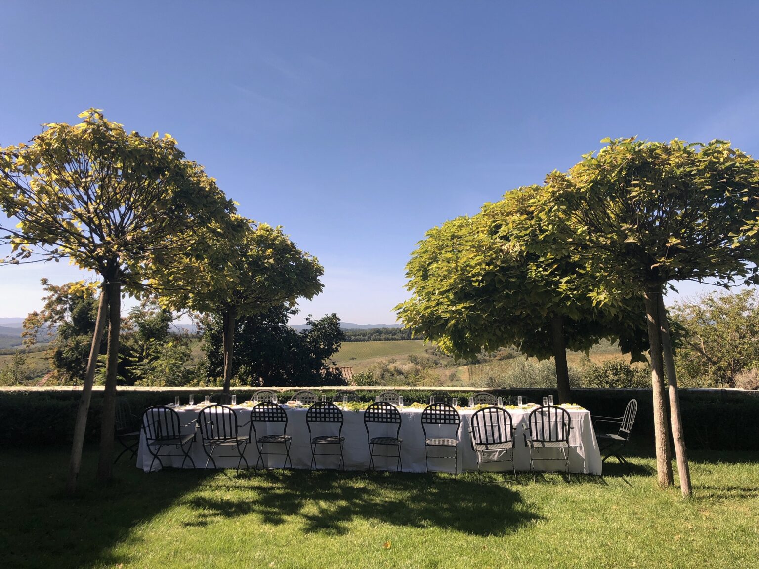 Outdoor dining setup under trees with a scenic view, featuring a long table covered with a white tablecloth and surrounded by chairs, ideal for gatherings or events in nature. | Only on SopranoVillas