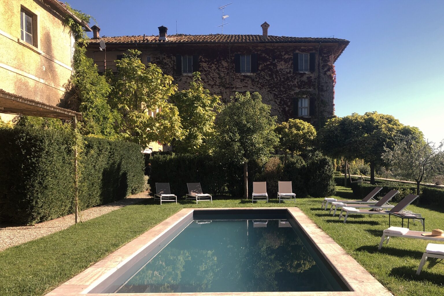 Scenic view of a tranquil swimming pool surrounded by lush greenery and lounge chairs, with a rustic villa in the background under a clear blue sky. Ideal for relaxation and leisure. | Only on SopranoVillas