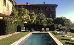 Scenic view of a tranquil swimming pool surrounded by lush greenery and lounge chairs, with a rustic villa in the background under a clear blue sky. Ideal for relaxation and leisure.