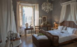 Cozy bedroom interior featuring a wooden bed, elegant chandelier, and large window with natural light. Decor includes a vintage table, decorative items, and soft linens, creating a warm and inviting atmosphere.