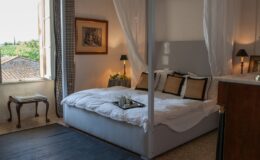Cozy bedroom featuring a four-poster bed with white drapes, elegant decor, and a scenic view through a window. The room includes a bedside table with lamps and a small seating area, creating a serene atmosphere.