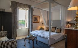 Elegant bedroom featuring a four-poster bed with white drapes, cozy seating area, and a window with scenic views. The decor combines classic and contemporary styles, creating a warm and inviting atmosphere. Ideal for relaxation and comfort.