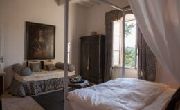 Cozy bedroom featuring a four-poster bed with white linens, a vintage painting on the wall, and a comfortable sitting area with plush cushions. Natural light streams through a large window, highlighting the elegant decor and rustic charm of the room.