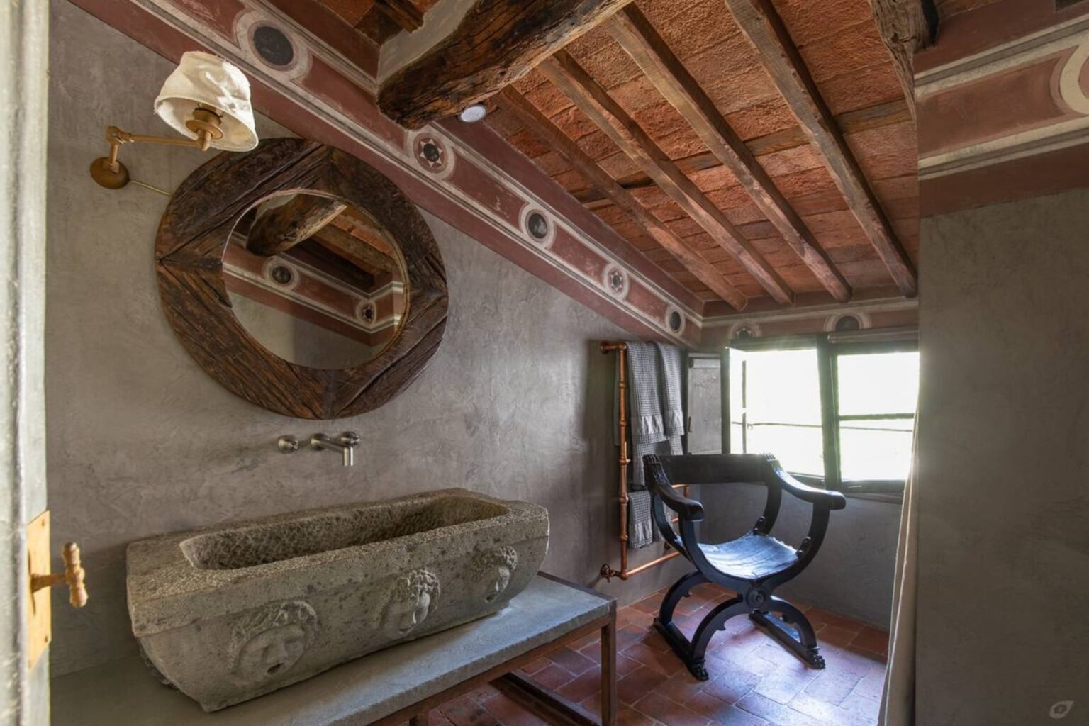 Interior view of a rustic bathroom featuring a stone sink, wooden chair, and a round mirror, with exposed wooden beams and natural light from a window. | Only on SopranoVillas