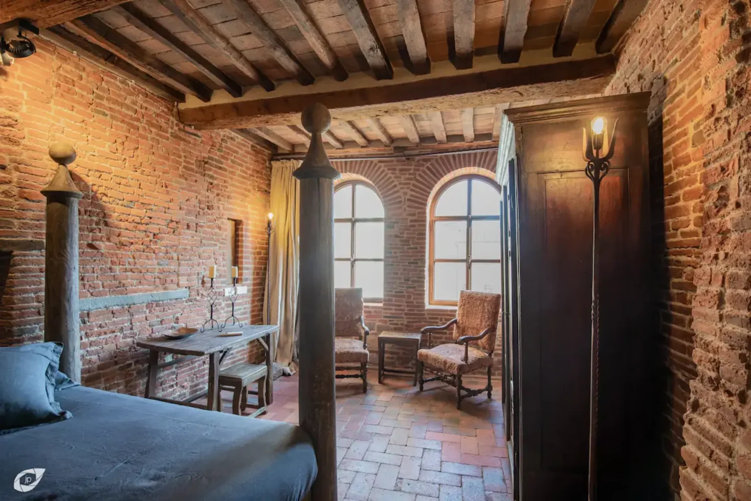 Cozy rustic bedroom featuring exposed brick walls, wooden beams, and large arched windows. The room includes a four-poster bed, a wooden wardrobe, and two vintage rocking chairs, creating a warm and inviting atmosphere. | Only on SopranoVillas