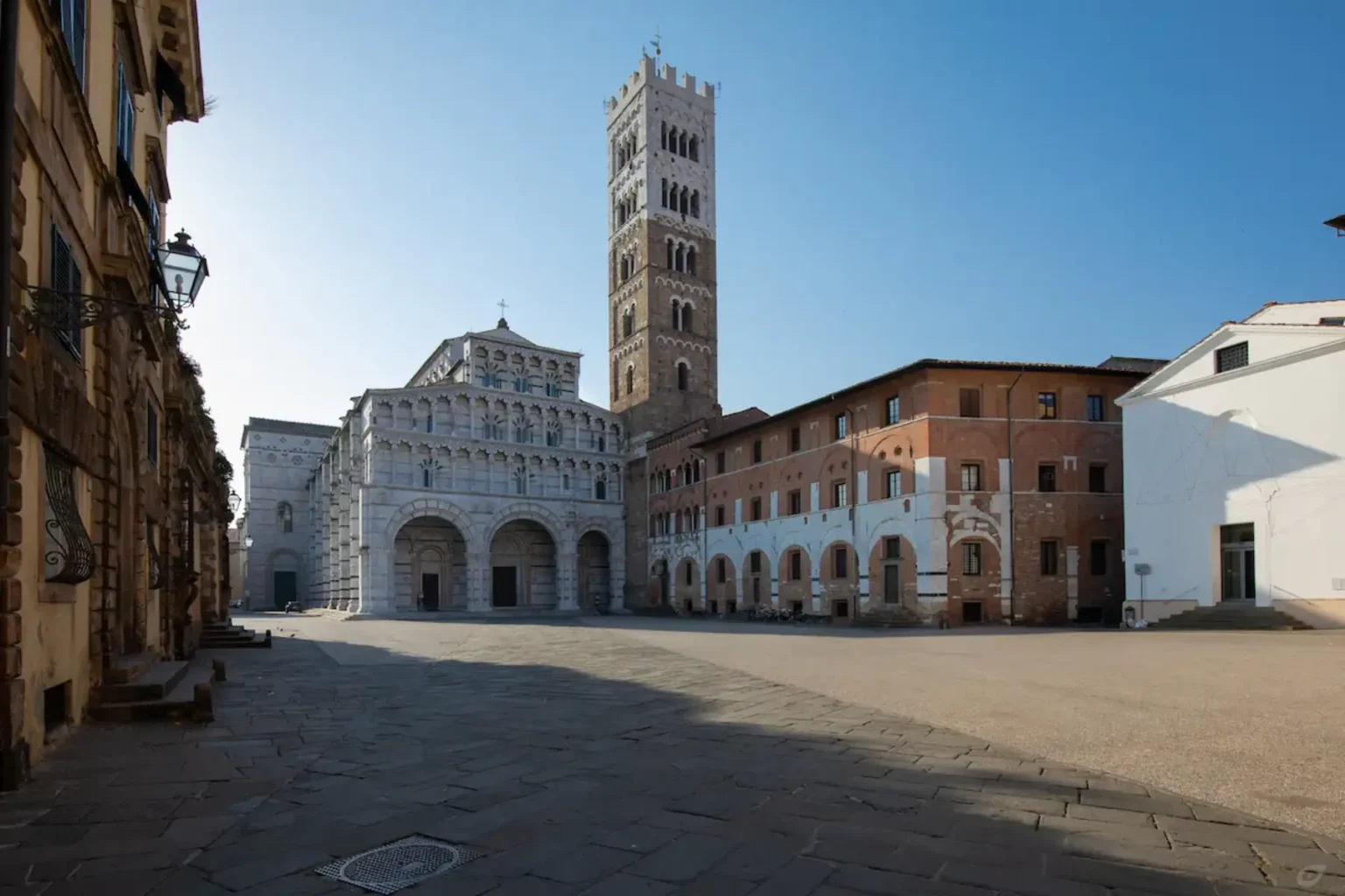 Historic architecture in Lucca, Italy, featuring the impressive Torre delle Ore and the intricate facade of the Basilica di San Frediano, set against a clear blue sky. | Only on SopranoVillas