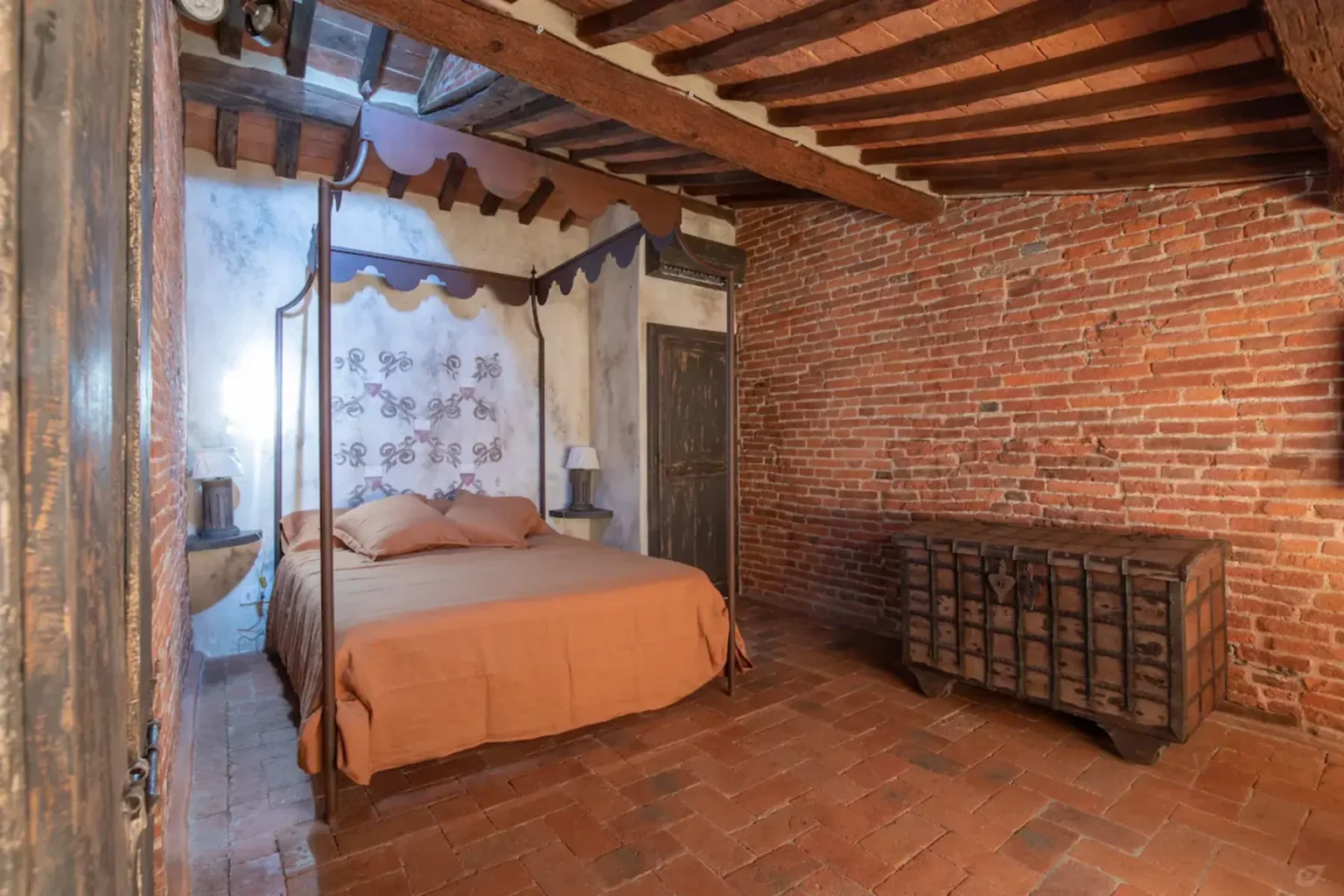 Cozy rustic bedroom featuring a four-poster bed with an orange bedspread, exposed brick walls, wooden beams, and a vintage wooden dresser, creating a warm and inviting atmosphere. | Only on SopranoVillas