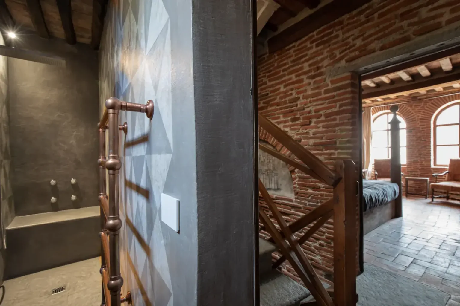 Interior view of a rustic home featuring a modern shower area with geometric wall design, a copper towel rack, and exposed brick walls leading to a staircase. Natural light filters through large windows in the adjoining room, highlighting wooden furniture and warm tones. | Only on SopranoVillas