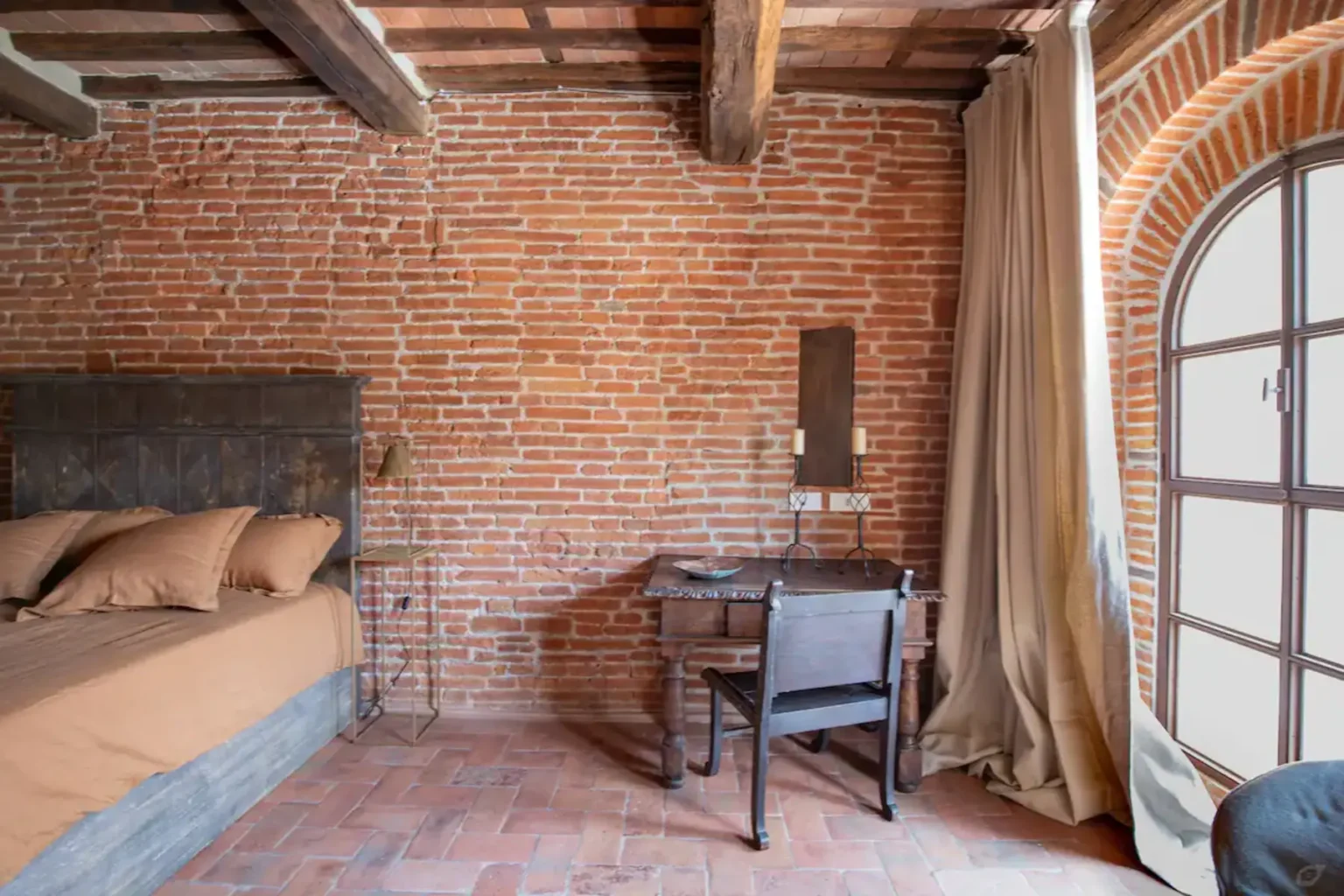Cozy bedroom featuring exposed brick walls, a wooden bed with beige linens, a vintage desk with a chair, and a large window with curtains, creating a warm and inviting atmosphere. | Only on SopranoVillas