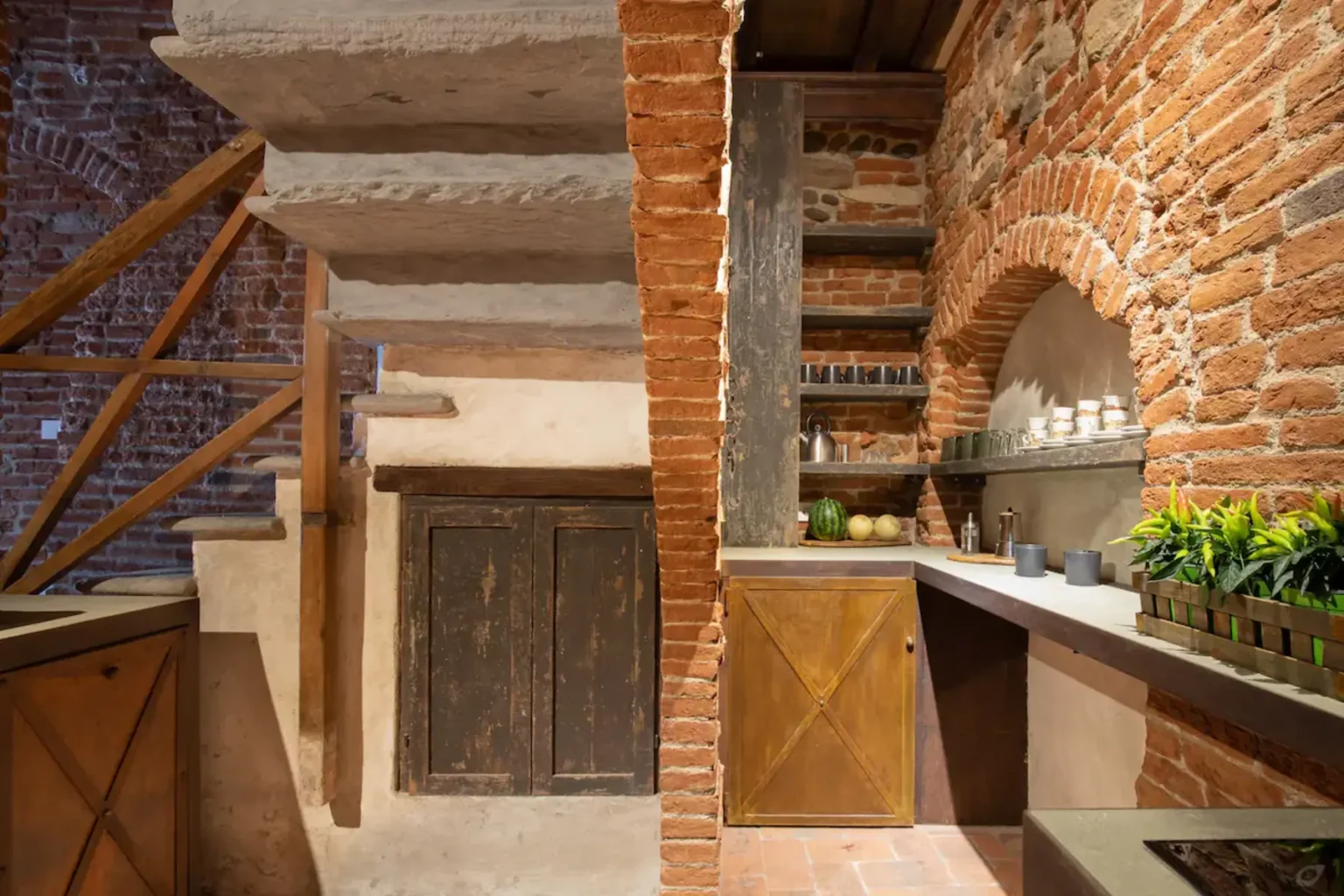 Modern kitchen design featuring exposed brick walls, a wooden staircase, and rustic cabinetry. The space includes a counter with plants and decorative items, blending contemporary and traditional elements for a cozy atmosphere. | Only on SopranoVillas