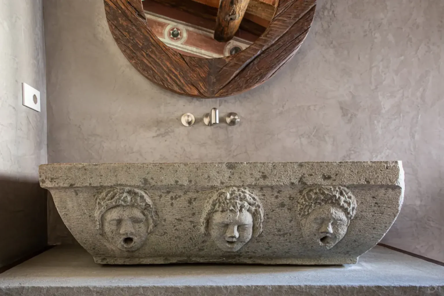 Stone sink with three carved faces, featuring intricate details, set against a textured wall and complemented by a wooden mirror above. Modern faucet fixtures complete the elegant bathroom design. | Only on SopranoVillas