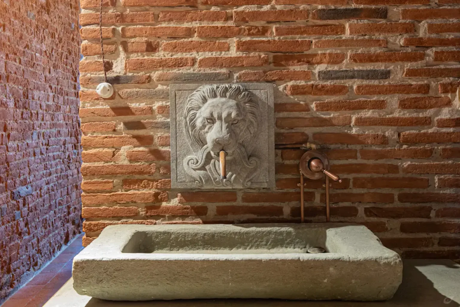 Stone lion fountain with water spout against a rustic brick wall, featuring a rectangular basin below for collecting water. | Only on SopranoVillas
