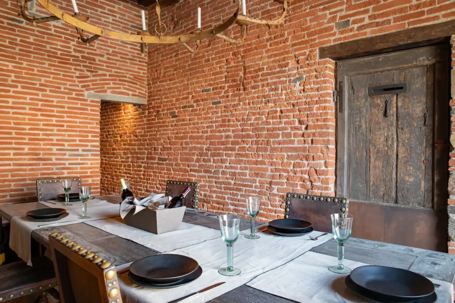 Dining room featuring a rustic wooden table set with black plates and glassware, surrounded by exposed red brick walls and a vintage wooden door. A centerpiece basket holds wine bottles, complemented by a chandelier with candles. | Only on SopranoVillas