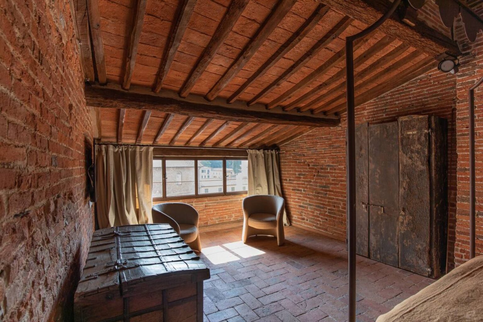 Cozy attic room featuring exposed brick walls, wooden beams, and natural light from a large window. Includes two comfortable chairs and a rustic wooden chest, creating a warm and inviting atmosphere. | Only on SopranoVillas