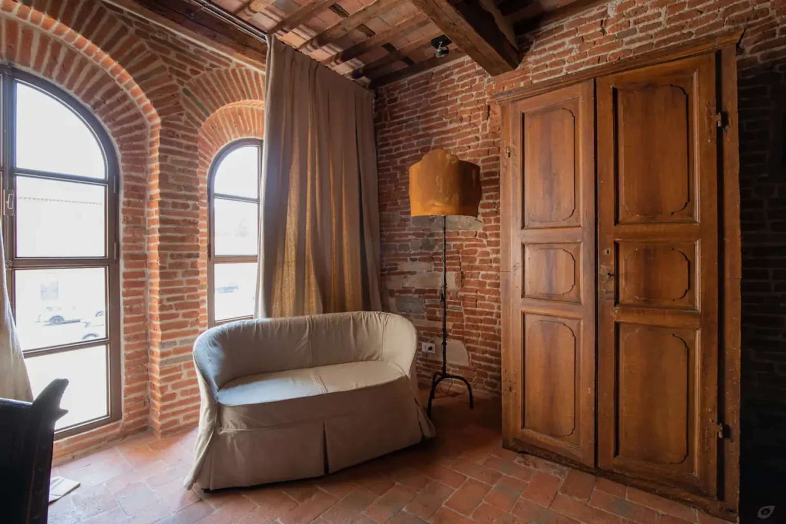 Cozy interior of a room featuring exposed brick walls, a comfortable sofa, a wooden wardrobe, and large arched windows that allow natural light to fill the space. | Only on SopranoVillas