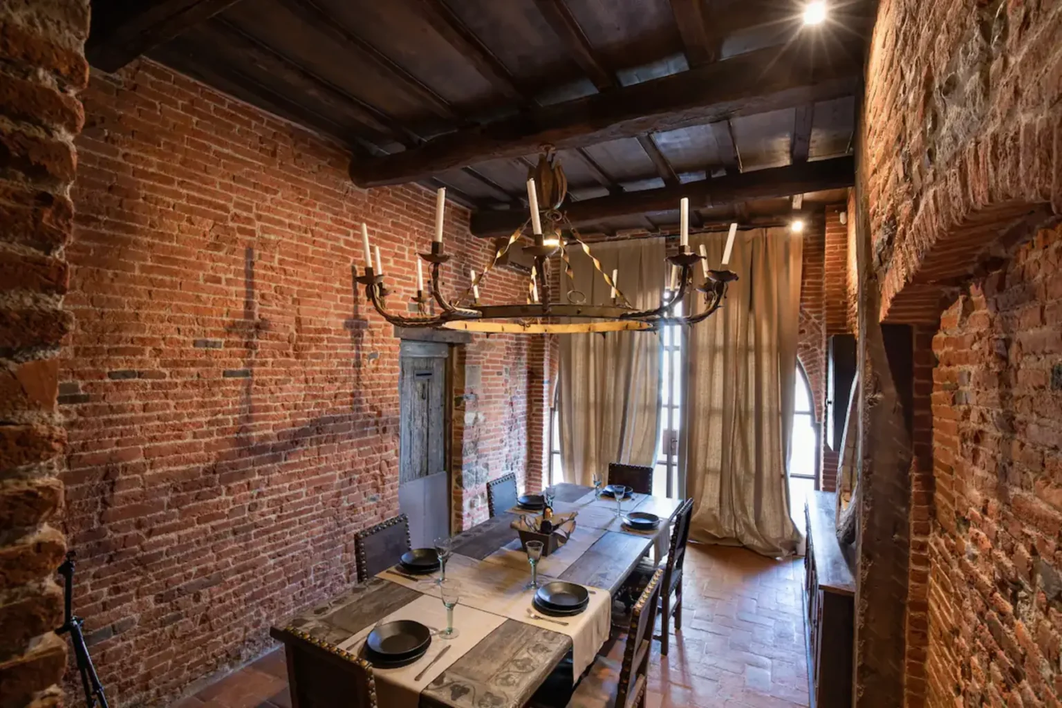 Rustic dining room featuring exposed brick walls, a wooden beam ceiling, and a vintage chandelier. A long dining table is set with plates and cutlery, illuminated by natural light from large windows draped with curtains. | Only on SopranoVillas