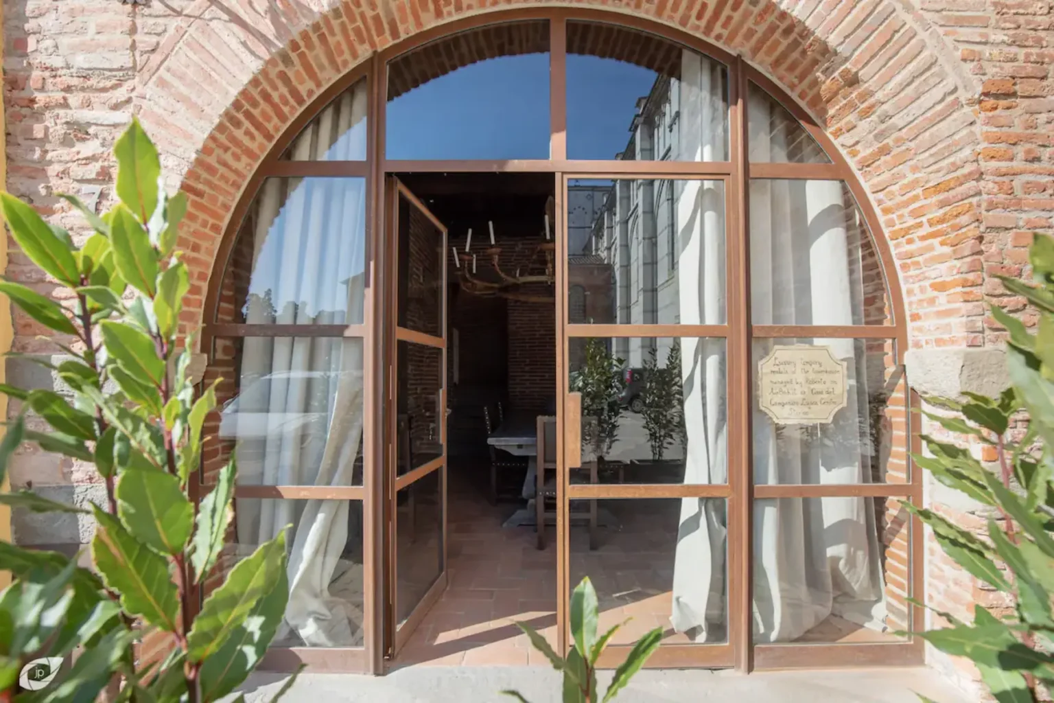 Entrance to a rustic restaurant featuring a large arched wooden door with glass panels, surrounded by potted plants and soft natural light. | Only on SopranoVillas
