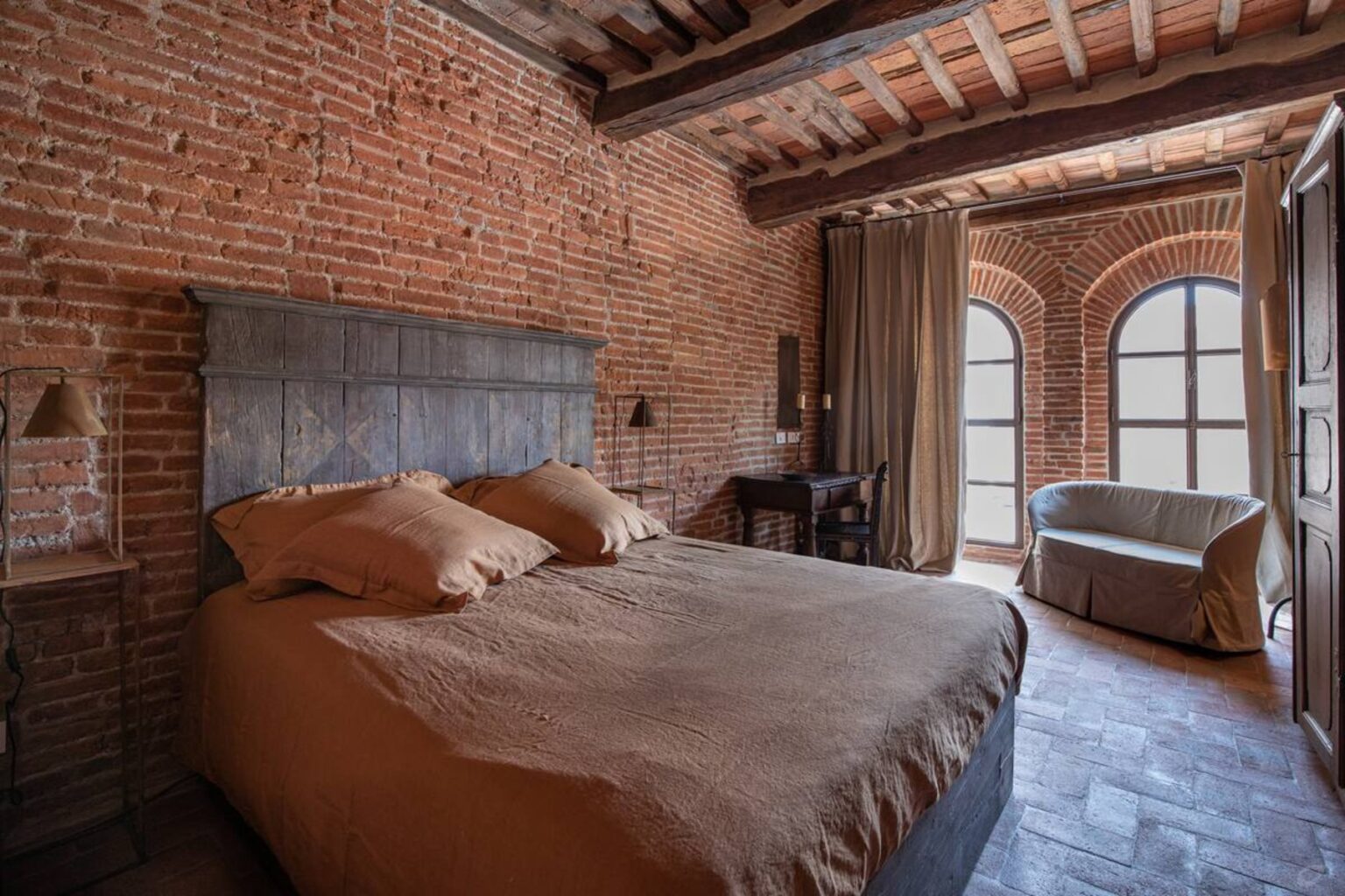 Cozy bedroom featuring a rustic design with exposed brick walls, a wooden ceiling, and a large bed with brown linens. Natural light streams through arched windows, illuminating a small sitting area with a couch. Ideal for a relaxing getaway or home decor inspiration. | Only on SopranoVillas