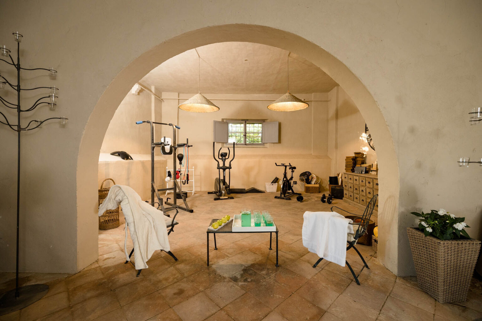 Home gym interior featuring exercise equipment, including an elliptical machine and stationary bikes, set in a spacious, well-lit room with a rustic design. The area includes a coffee table with refreshments, comfortable seating, and decorative plants, creating a welcoming atmosphere for workouts. | Only on SopranoVillas