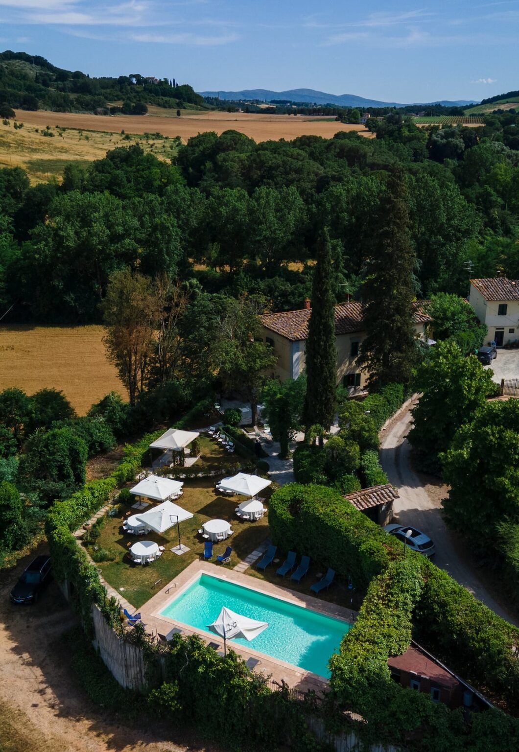 Scenic aerial view of a countryside villa featuring a turquoise swimming pool, surrounded by lush greenery and sun loungers, with outdoor seating areas under white umbrellas. The landscape includes fields and distant hills, creating a peaceful retreat atmosphere. | Only on SopranoVillas