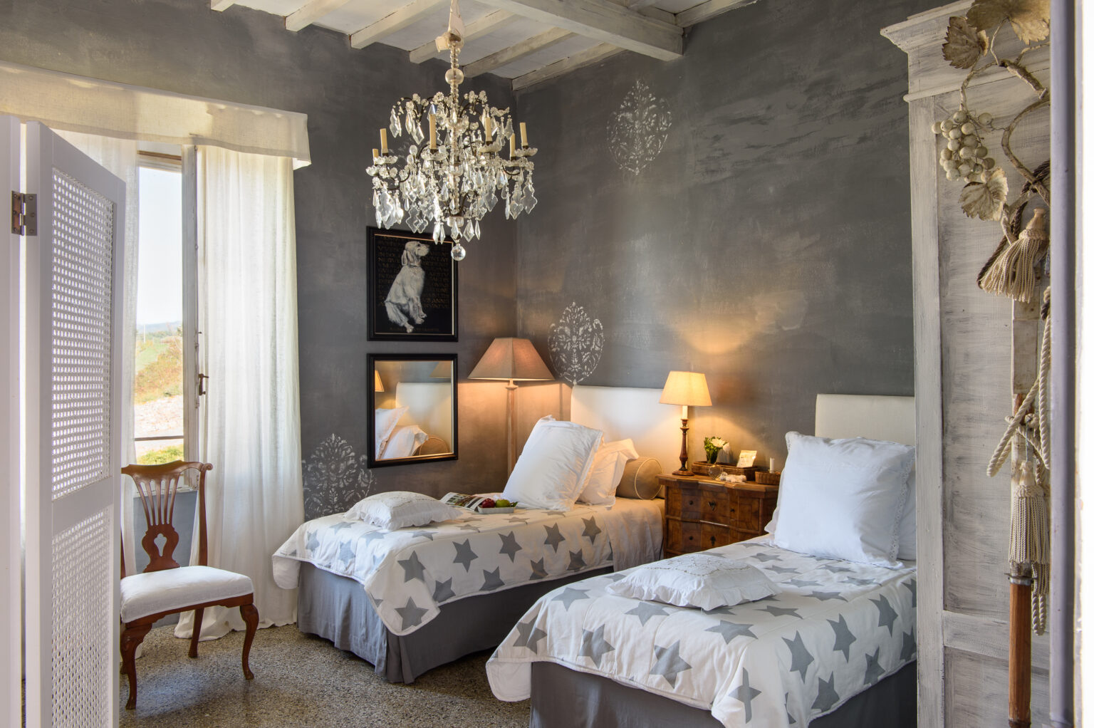 Cozy bedroom featuring two twin beds with star-patterned bedding, a vintage chandelier, and elegant decor. Natural light streams in through large windows, enhancing the inviting atmosphere. Ideal for relaxation and comfort. | Only on SopranoVillas