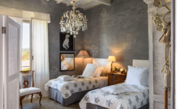 Cozy bedroom featuring two twin beds with star-patterned bedding, a vintage chandelier, and elegant decor. Natural light streams in through large windows, enhancing the inviting atmosphere. Ideal for relaxation and comfort.