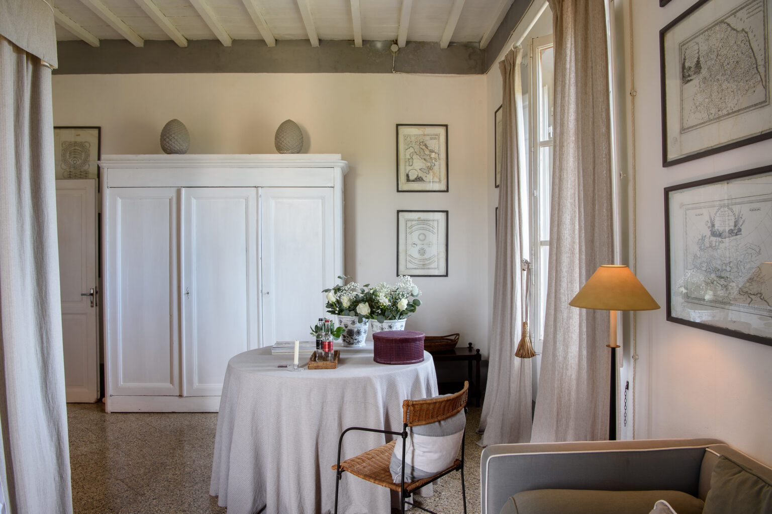 Bright and airy living space featuring a round table adorned with flowers, a cozy chair, and a modern lamp. The room has white walls, a wooden ceiling, and framed artwork, creating a stylish and inviting atmosphere. | Only on SopranoVillas