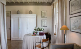 Bright and airy living space featuring a round table adorned with flowers, a cozy chair, and a modern lamp. The room has white walls, a wooden ceiling, and framed artwork, creating a stylish and inviting atmosphere.