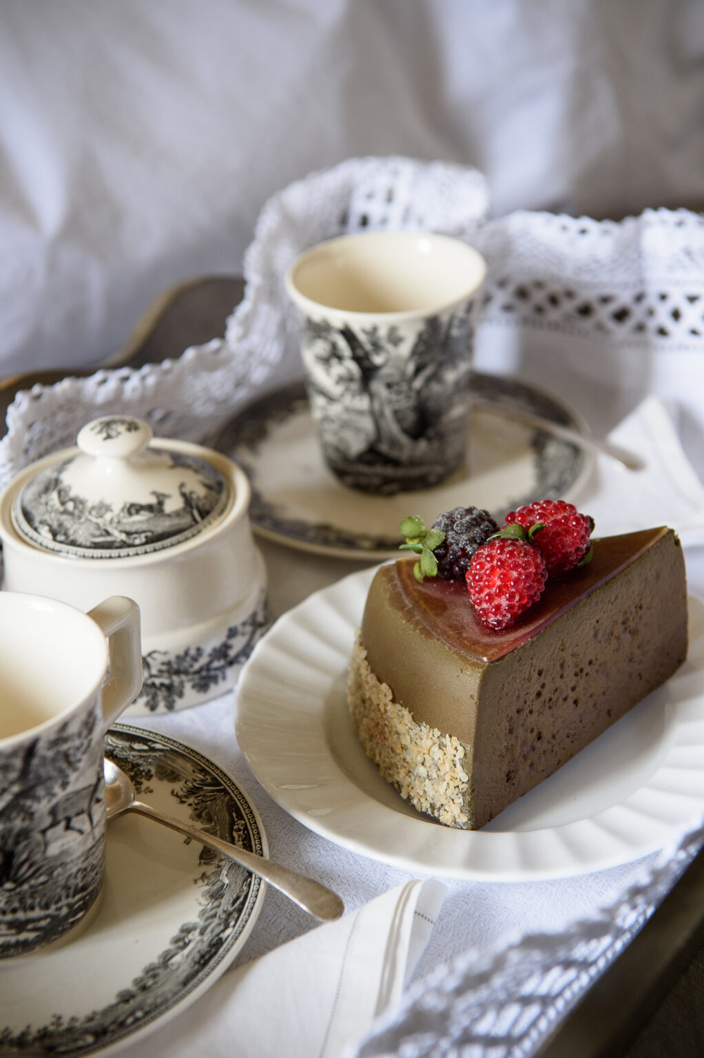 A beautifully arranged dessert table featuring a slice of chocolate mousse cake topped with fresh raspberries and blackberries, accompanied by vintage porcelain dishes, a teacup, and a sugar bowl, all set on a delicate lace tablecloth. | Only on SopranoVillas