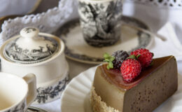 A beautifully arranged dessert table featuring a slice of chocolate mousse cake topped with fresh raspberries and blackberries, accompanied by vintage porcelain dishes, a teacup, and a sugar bowl, all set on a delicate lace tablecloth.