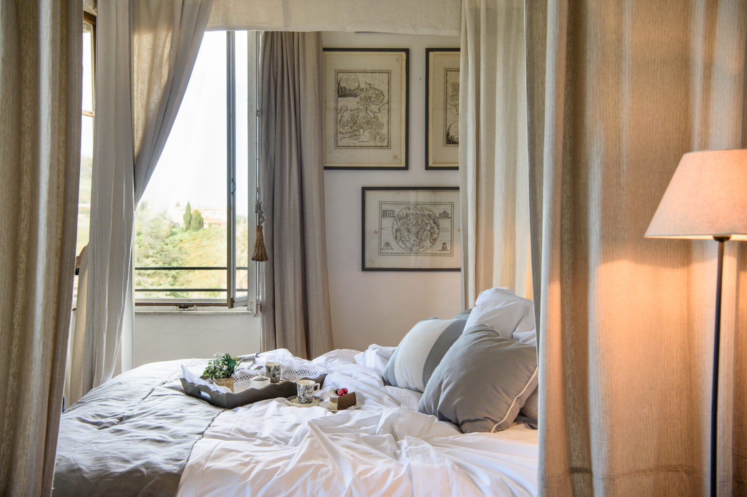 Cozy bedroom featuring a neatly made bed with gray and white bedding, decorative pillows, and a tray with tea and snacks. Natural light streams in through a window adorned with light curtains, while vintage maps hang on the wall, creating a serene and inviting atmosphere. | Only on SopranoVillas