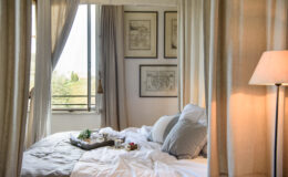 Cozy bedroom featuring a neatly made bed with gray and white bedding, decorative pillows, and a tray with tea and snacks. Natural light streams in through a window adorned with light curtains, while vintage maps hang on the wall, creating a serene and inviting atmosphere.