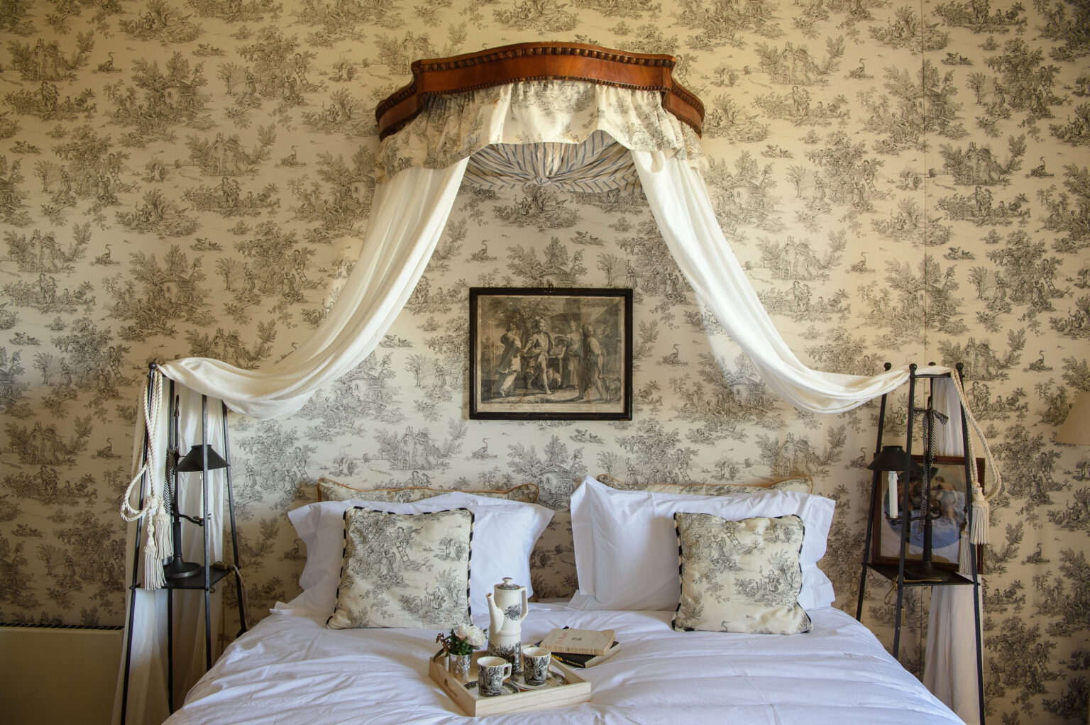 Elegant bedroom featuring a vintage canopy bed with sheer drapes, adorned with decorative pillows and a floral wallpaper backdrop. A framed artwork hangs above the bed, enhancing the room's classic charm. | Only on SopranoVillas