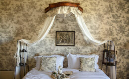 Elegant bedroom featuring a vintage canopy bed with sheer drapes, adorned with decorative pillows and a floral wallpaper backdrop. A framed artwork hangs above the bed, enhancing the room's classic charm.