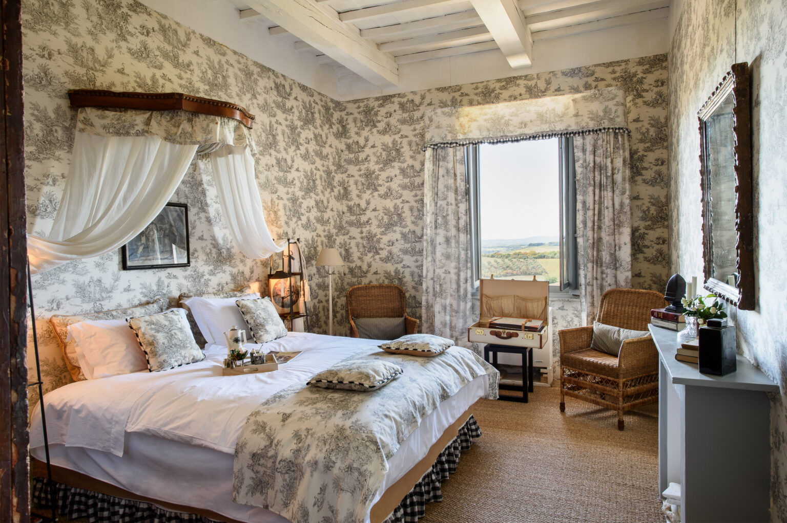 Cozy bedroom with floral wallpaper, a large bed adorned with decorative pillows, and wicker chairs by the window offering scenic views. Natural light illuminates the space, creating a warm and inviting atmosphere. | Only on SopranoVillas