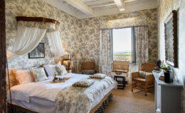 Cozy bedroom with floral wallpaper, a large bed adorned with decorative pillows, and wicker chairs by the window offering scenic views. Natural light illuminates the space, creating a warm and inviting atmosphere.