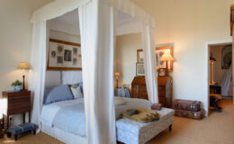 Spacious bedroom featuring a four-poster bed draped with white fabric, soft blue bedding, vintage decor, and warm lighting from bedside lamps, creating a cozy and inviting atmosphere.
