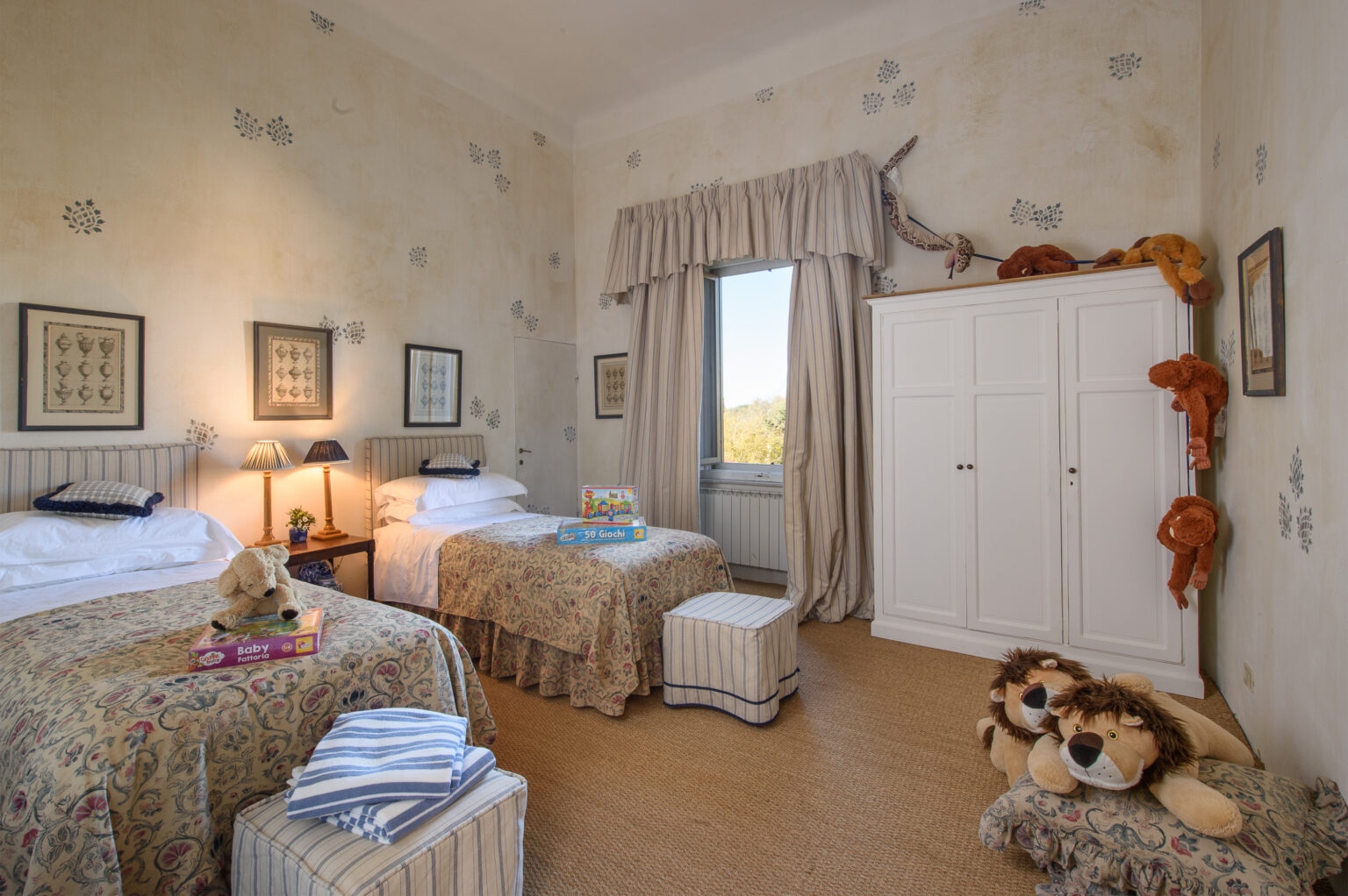 Cozy children's bedroom featuring two twin beds with floral bedding, a white wardrobe, and playful stuffed lions. Natural light streams through a window, enhancing the cheerful decor of the room. | Only on SopranoVillas