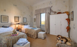 Cozy children's bedroom featuring two twin beds with floral bedding, a white wardrobe, and playful stuffed lions. Natural light streams through a window, enhancing the cheerful decor of the room.